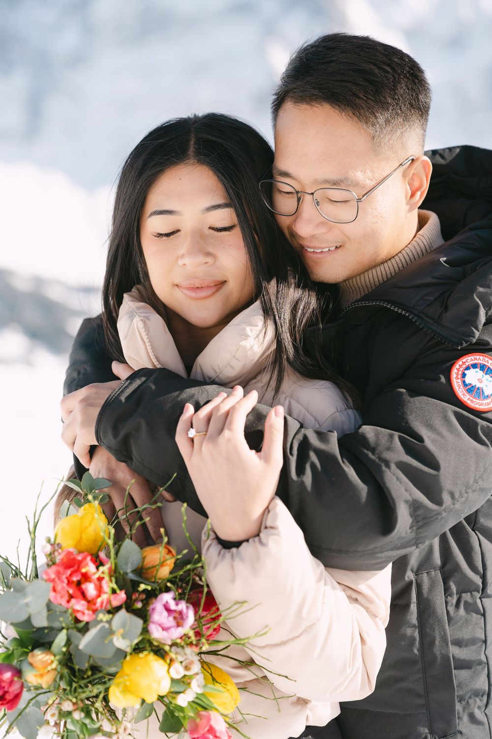 A romantic winter photoshoot featuring a couple in warm coats embracing while holding a colorful bouquet of flowers.