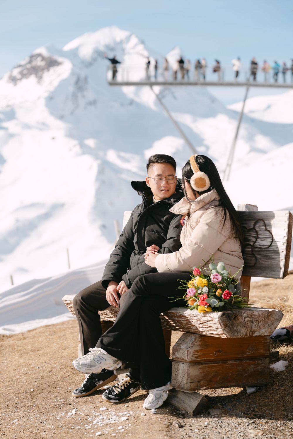 A couple enjoying a winter day on a mountain bench with snow-capped peaks and people in the background.