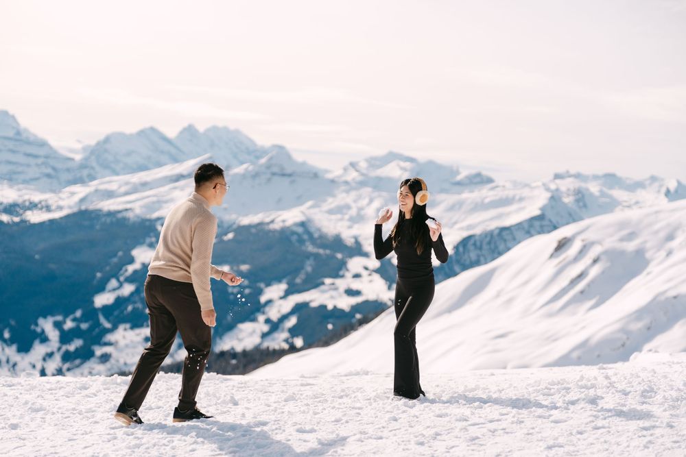 Skiers carve through fresh powder snow on a scenic mountain slope with snow-capped peaks in the background.