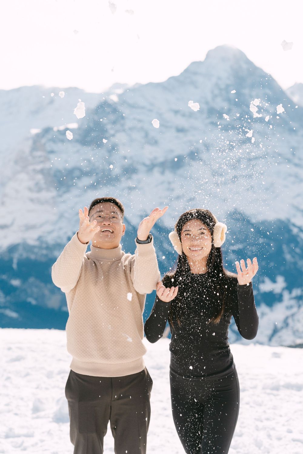 A playful couple throws snow in the air while enjoying a winter day in the mountains with snowy peaks in background.