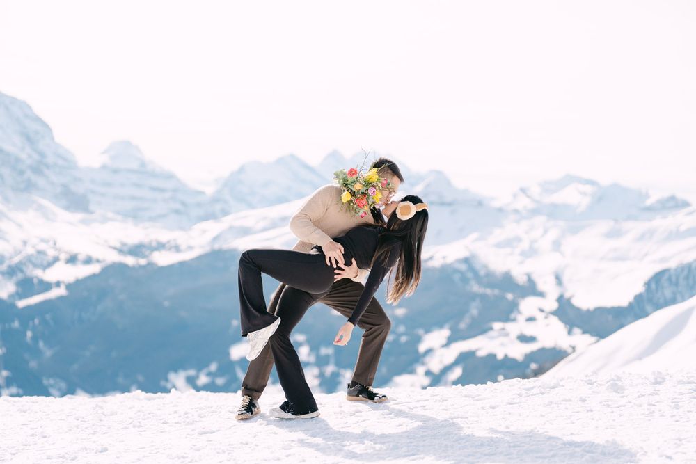 A couple in winter attire dances gracefully in the snow with majestic snow-capped mountains in the background.