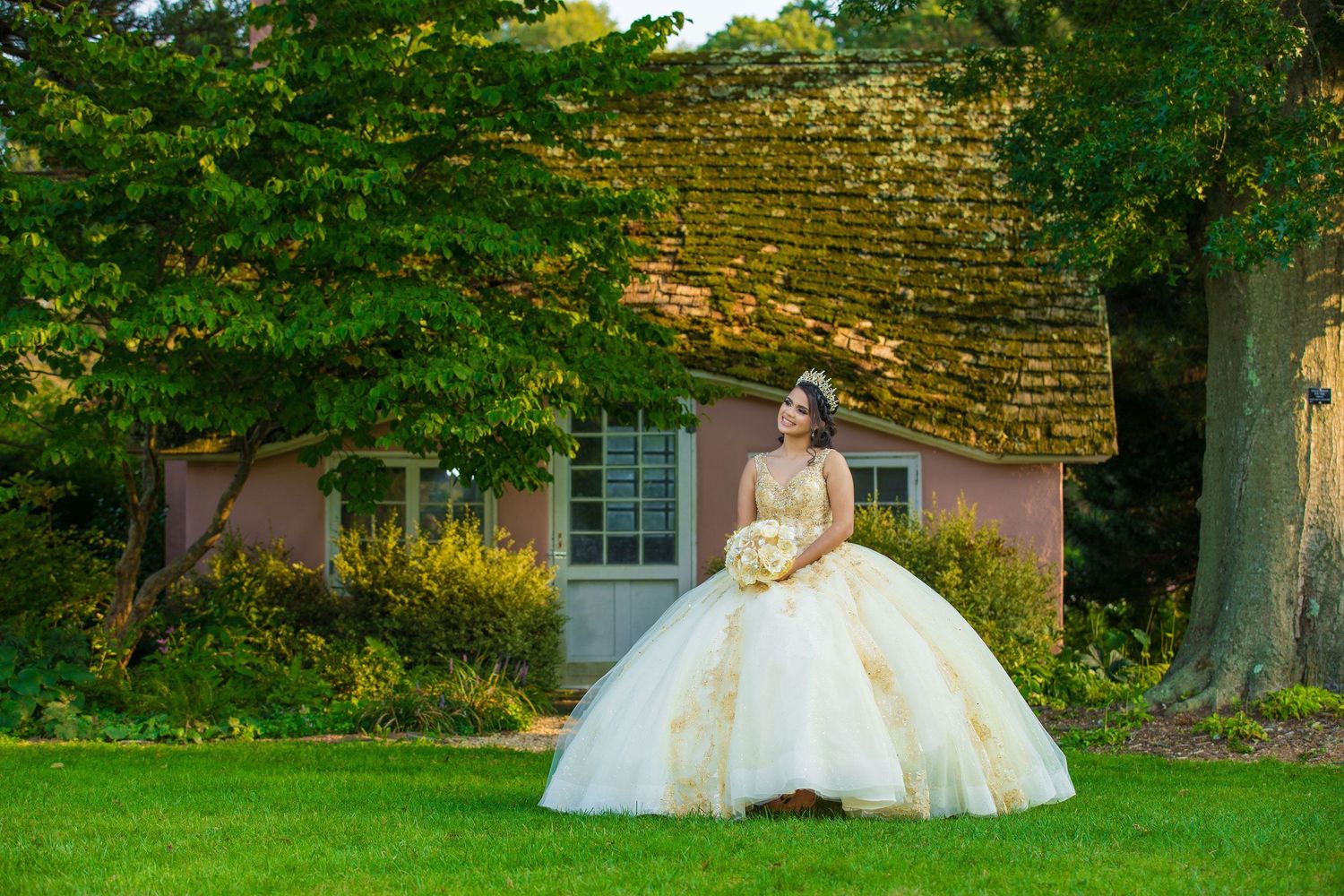 A voluminous cream-colored ballgown against a pink cottage with climbing vines and a stone wall in the background.