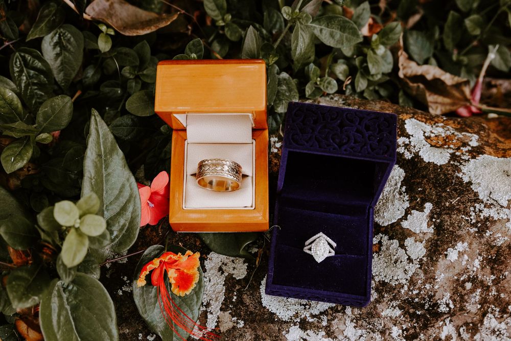 Two wedding rings displayed in orange and navy velvet ring boxes surrounded by tropical flowers.