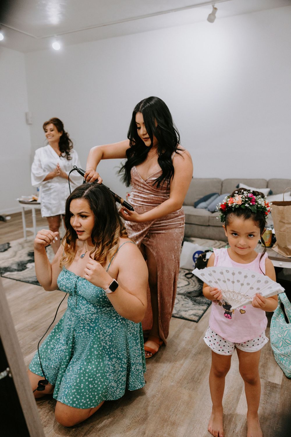 Bridesmaid curling hair of woman in teal dress while young flower girl watches in bright room.