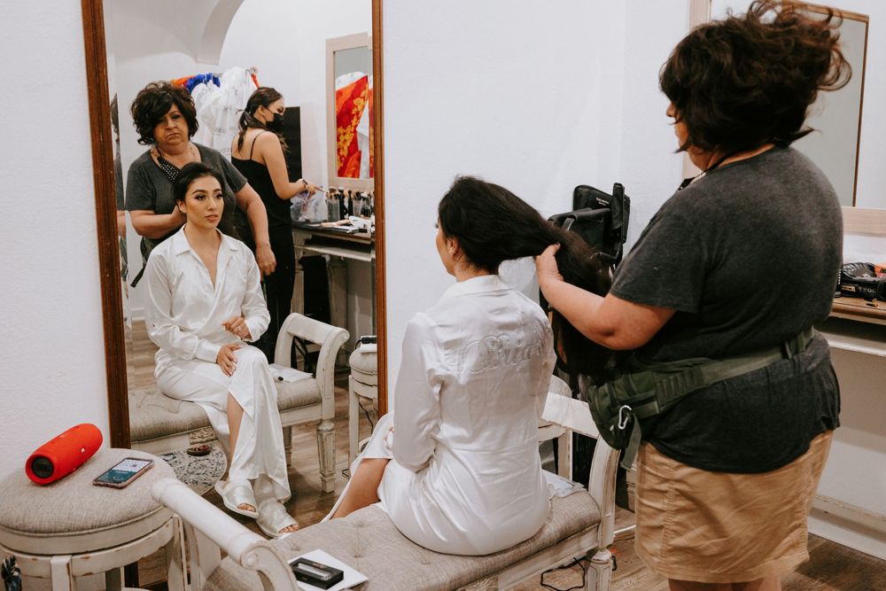 Bride in white robe getting hair and makeup done while videographer films bridal preparations in mirror.