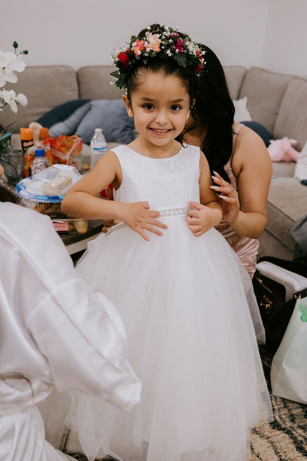 Adorable flower girl in white tulle dress and floral crown smiling during wedding preparations.