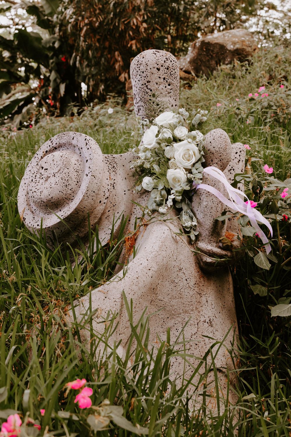 Stone garden statue holding white rose bouquet surrounded by pink flowers and green foliage.