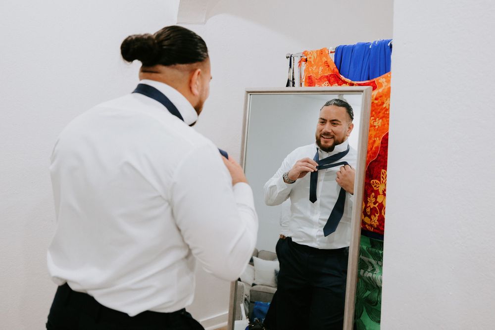 Groom tying navy blue necktie while looking in mirror during wedding day preparations.