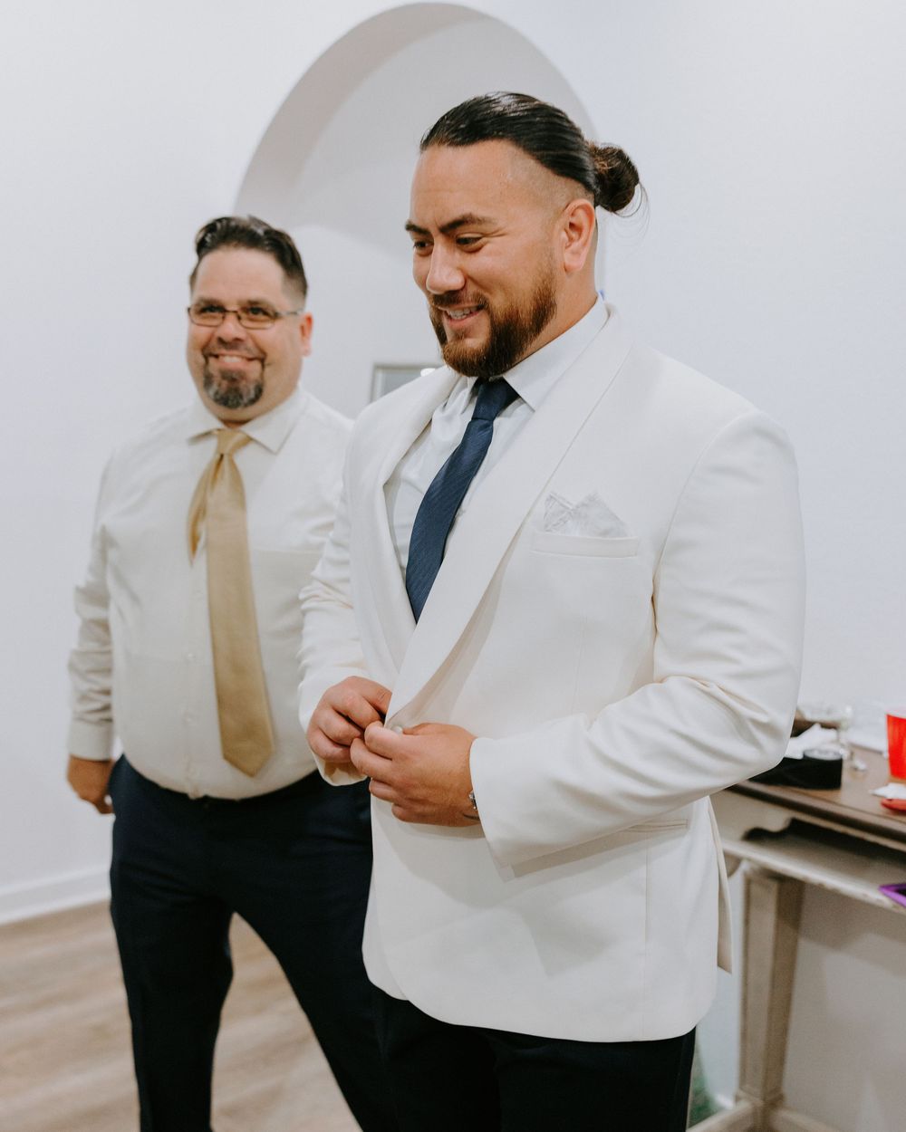 Groom buttoning white tuxedo jacket with groomsman in background during wedding preparations.