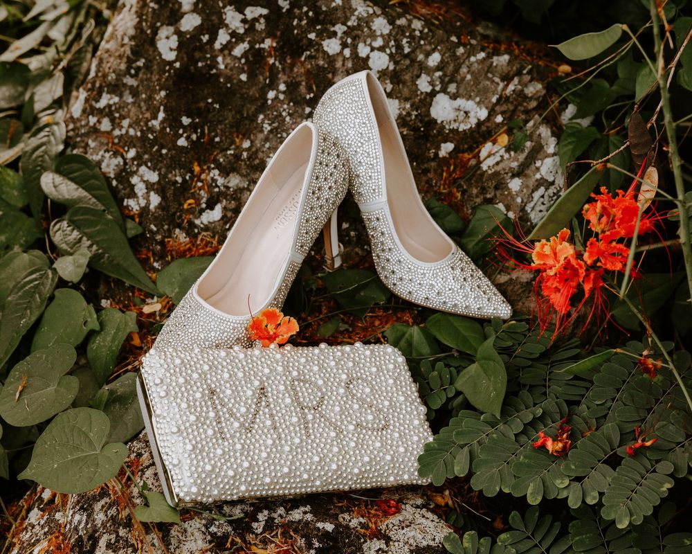 White pearl-embellished bridal heels and MRS clutch purse surrounded by tropical greenery and orange flowers.