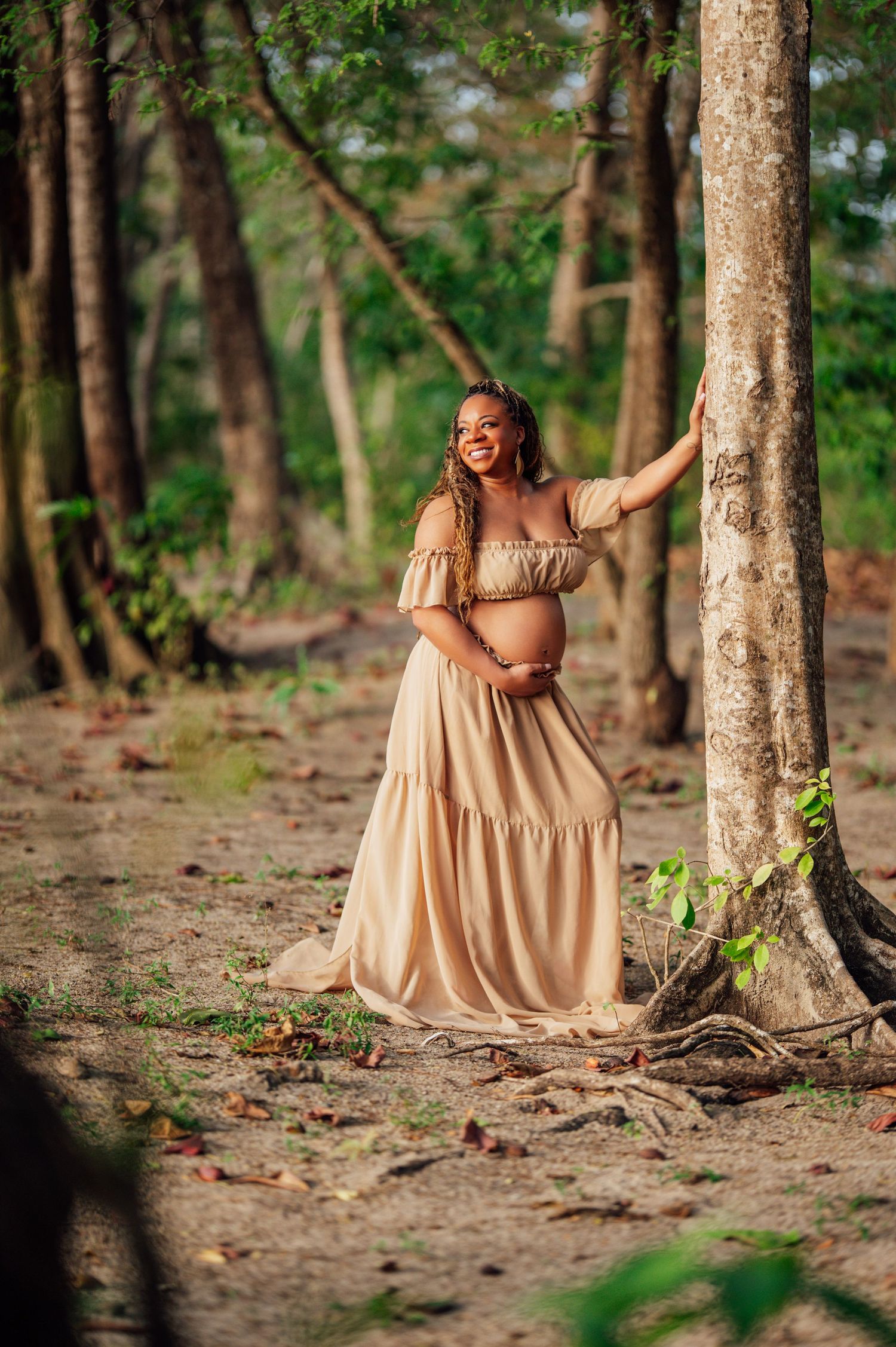A person in a flowing beige dress poses gracefully in a wooded setting with dappled sunlight filtering through trees.