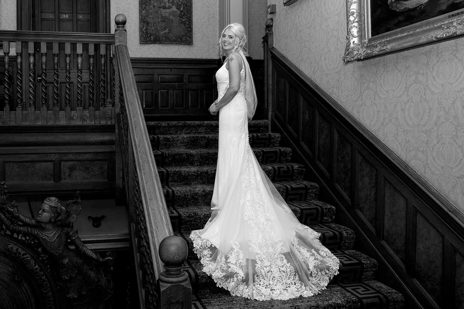 A bride in a flowing lace wedding gown stands elegantly on a grand wooden staircase in a historic venue at Armathwaite Hall in the Lakes
