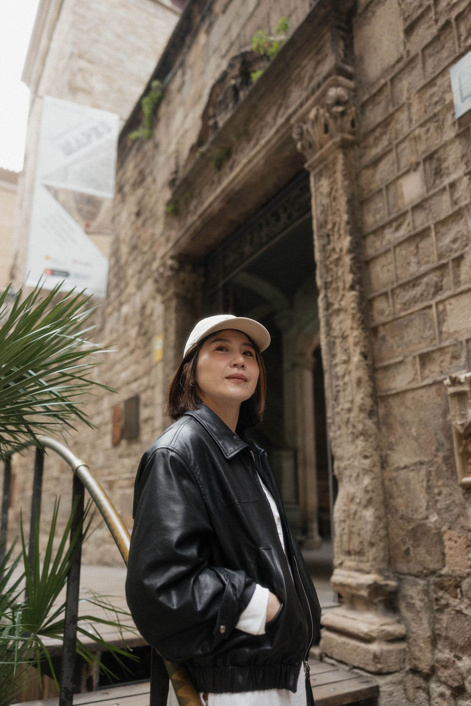 Young woman in leather jacket and polka dot skirt sitting on steps before Gothic cathedral in Barcelona.