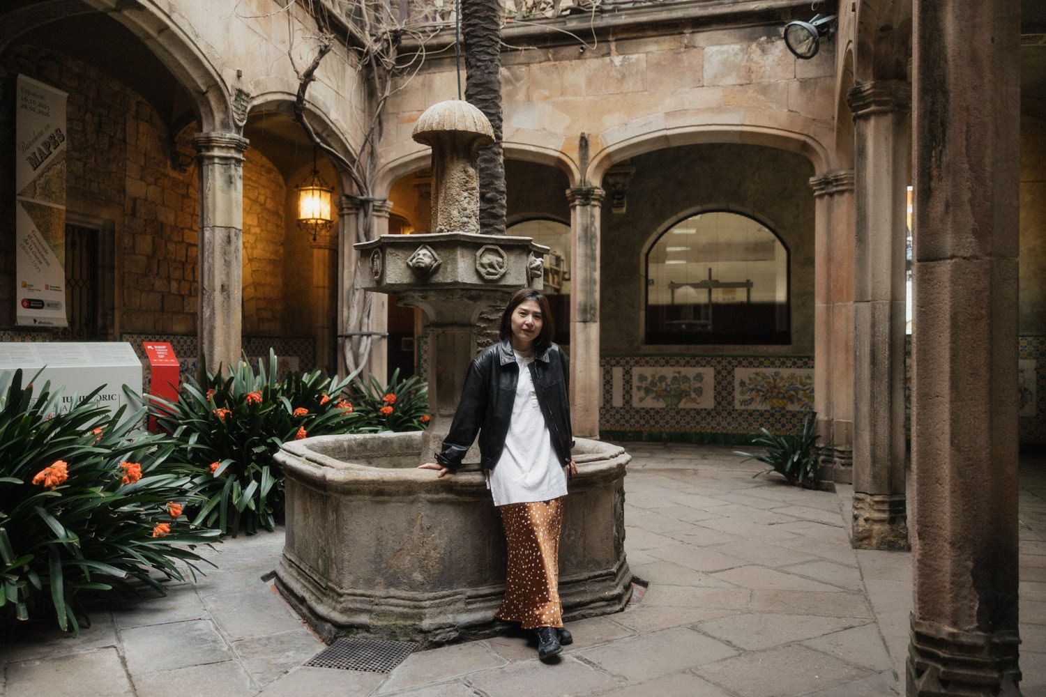 Woman in white dress and leather jacket poses by stone fountain in historic Gothic courtyard with arched architecture.