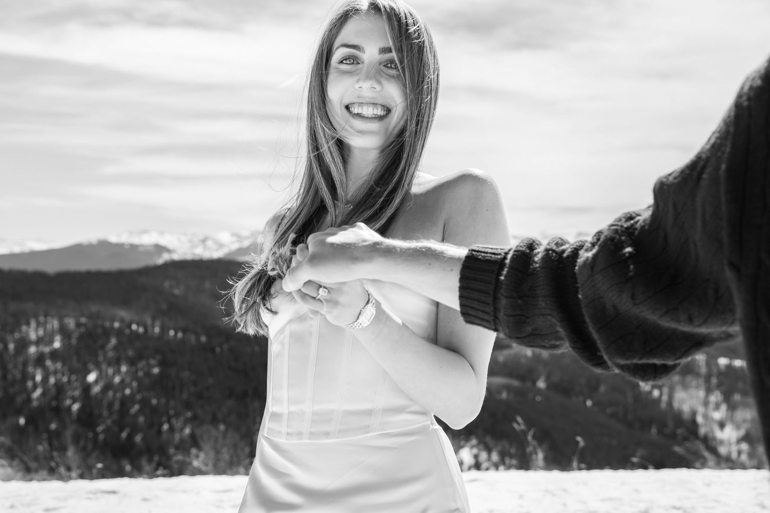 Black and white photo of a smiling woman holding hands with someone in a snowy mountain landscape for Vail Colorado engagement.