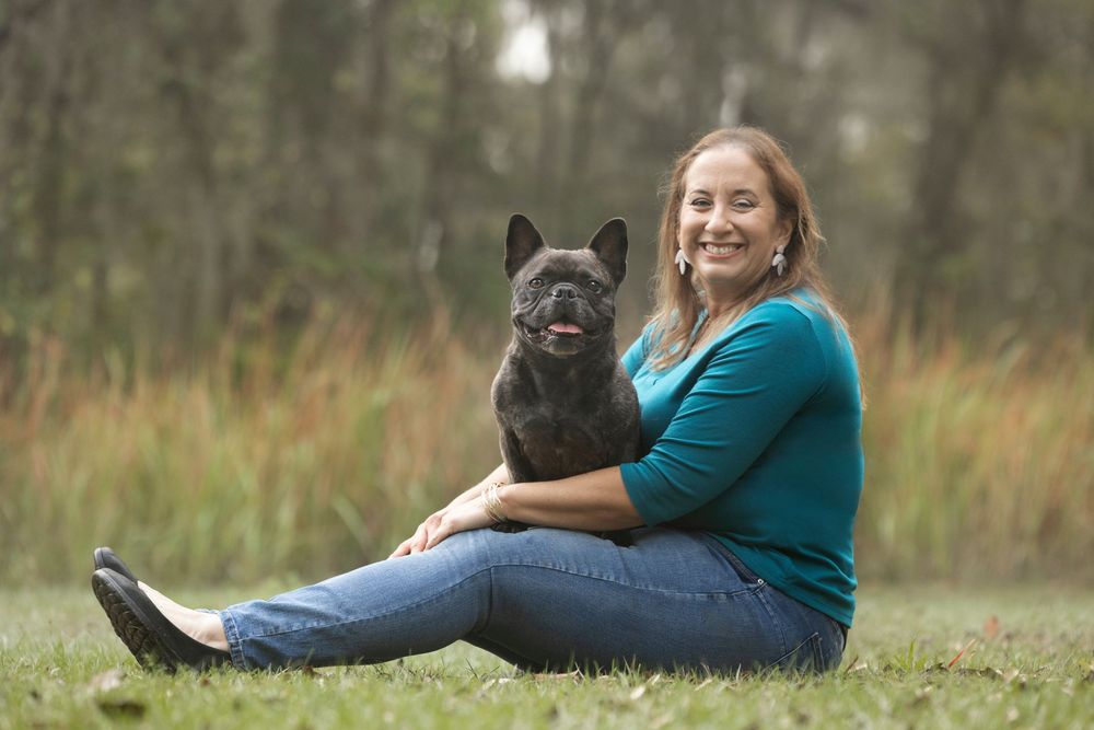 A woman sitting on a park lawn with her Frenchie resting comfortably on her lap.