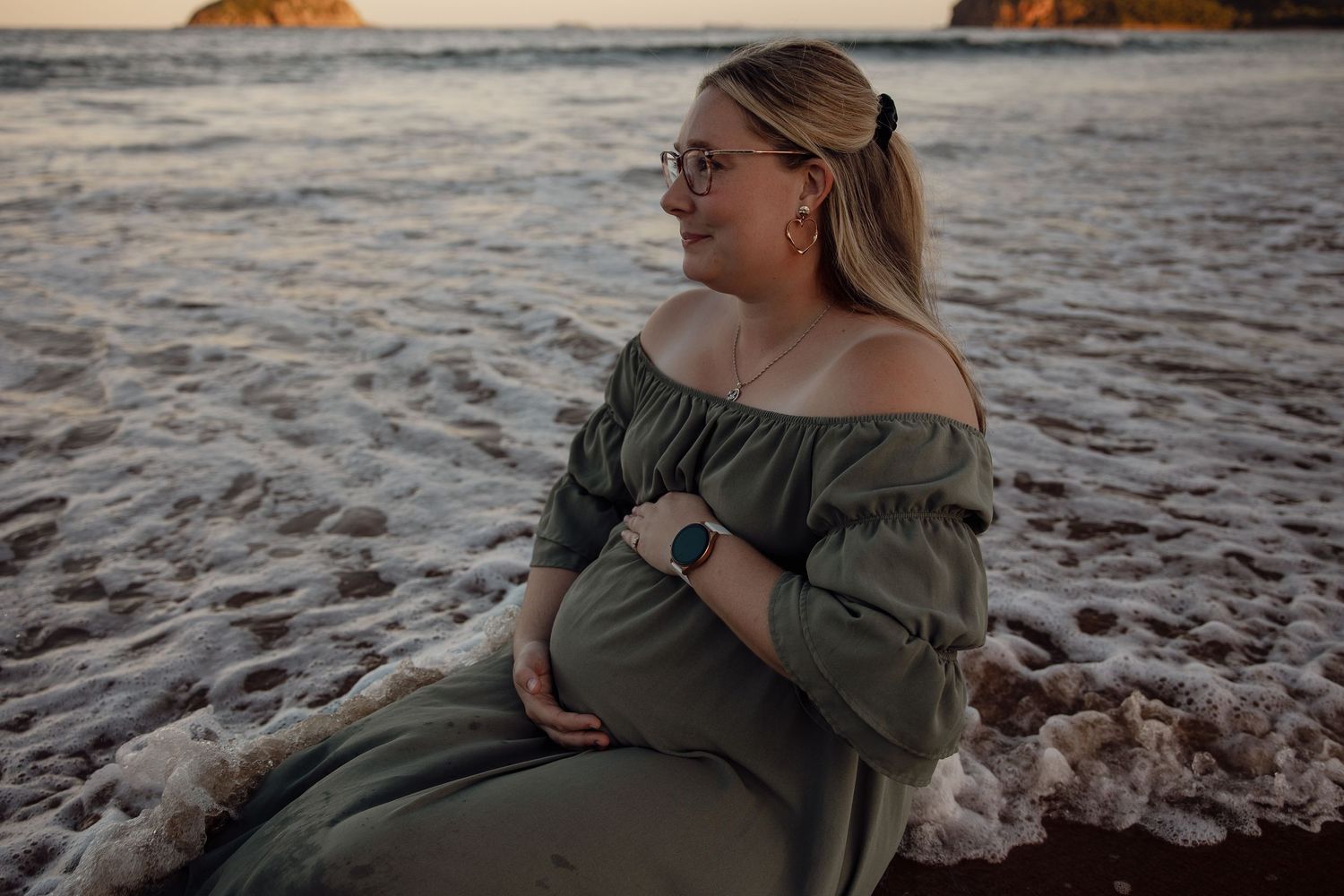 Pregnant woman in olive off-shoulder dress sitting on beach at sunset, cradling baby bump near ocean waves.