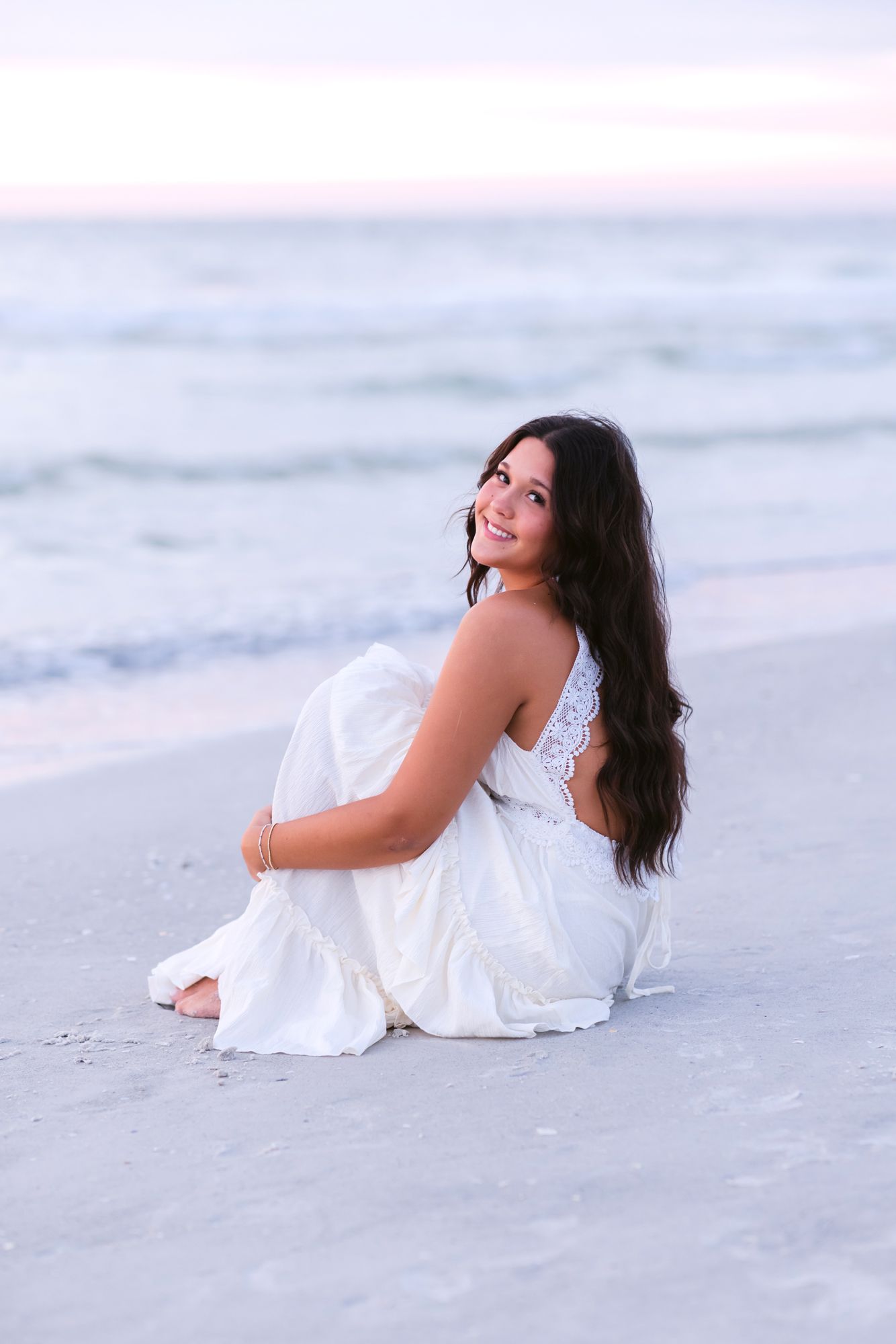 Dark, long haired senior girl wears a white dress sits with arms holding knees on Indian Rocks Beach at sunset in Clearwater, Florida.