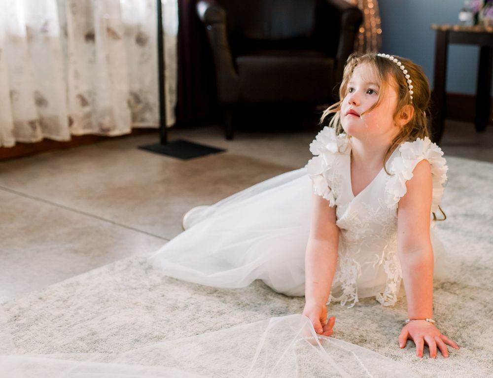 Bride in white lace dress kneels on carpet during wedding preparations.
