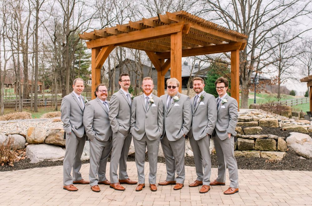 Groomsmen in matching gray suits lined up under wooden pergola for formal photo.