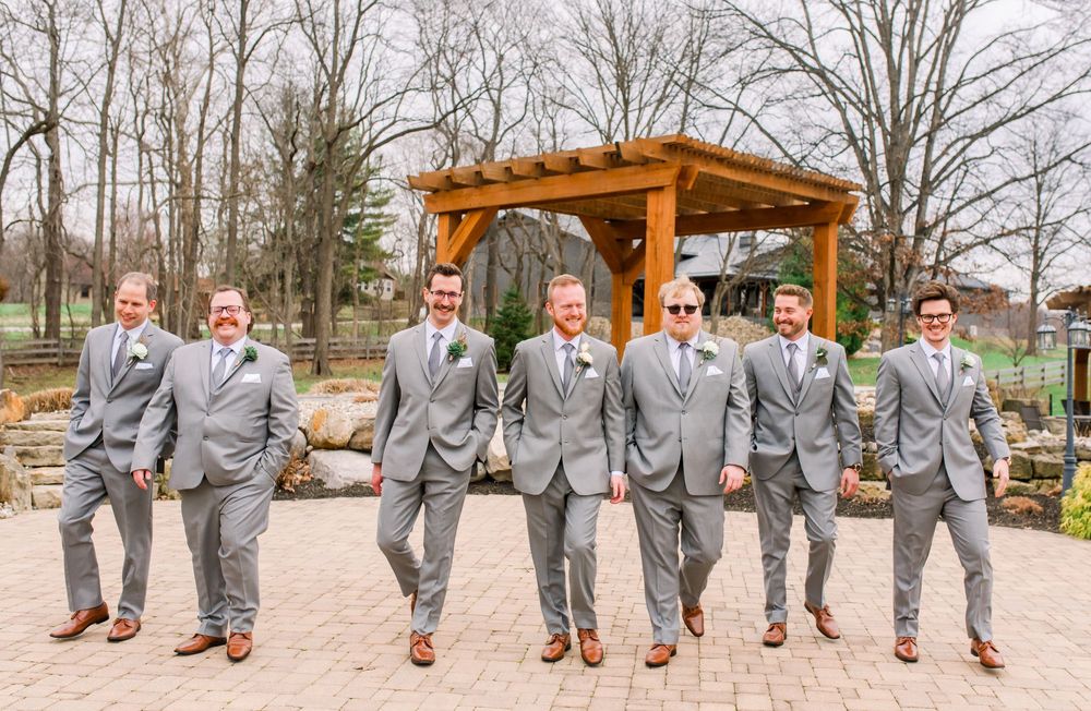 A row of groomsmen in gray suits show off their brown leather shoes in a coordinated outdoor photo.