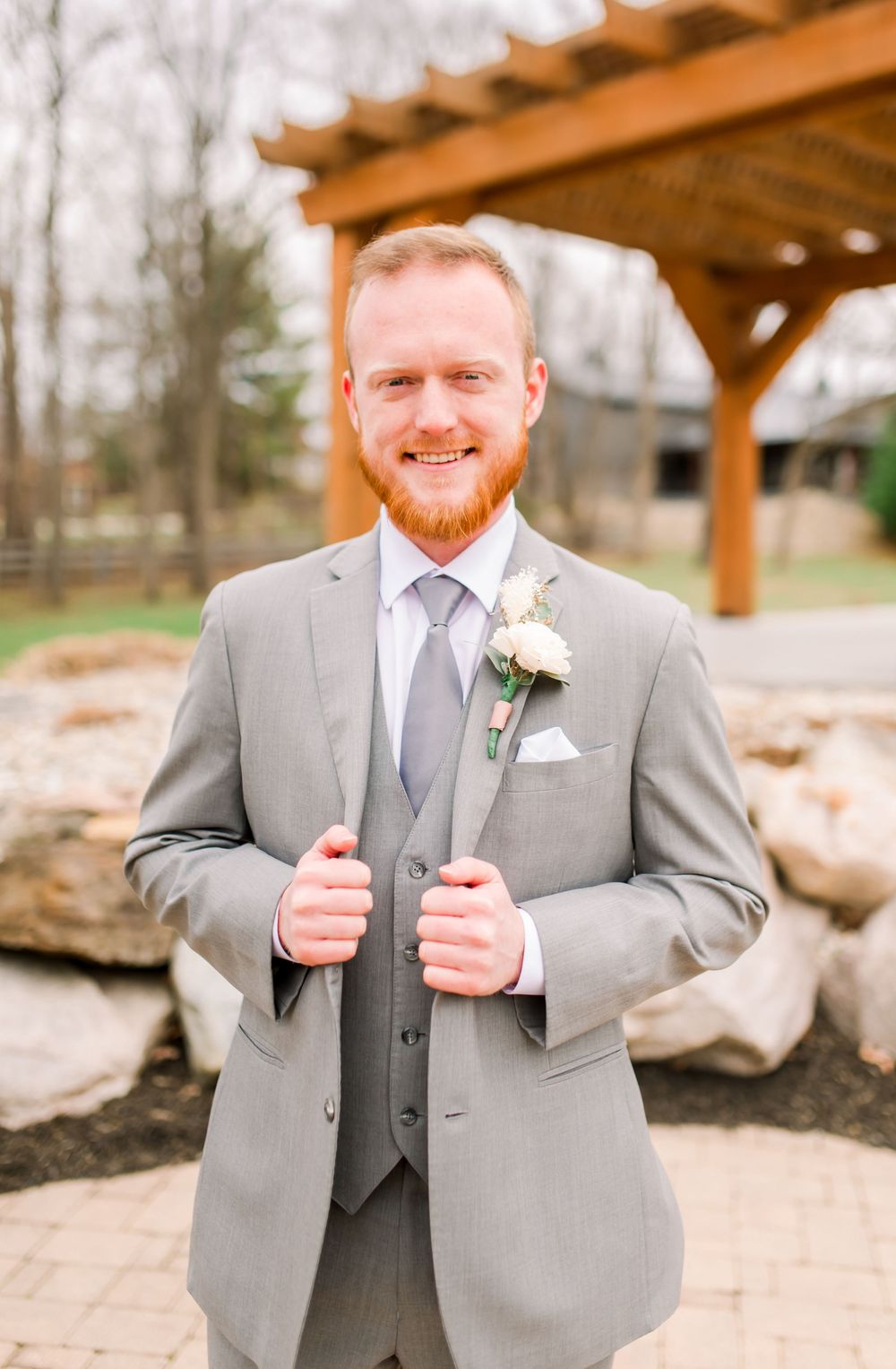 A groom in a light grey three-piece suit with boutonniere poses under a wooden pergola.