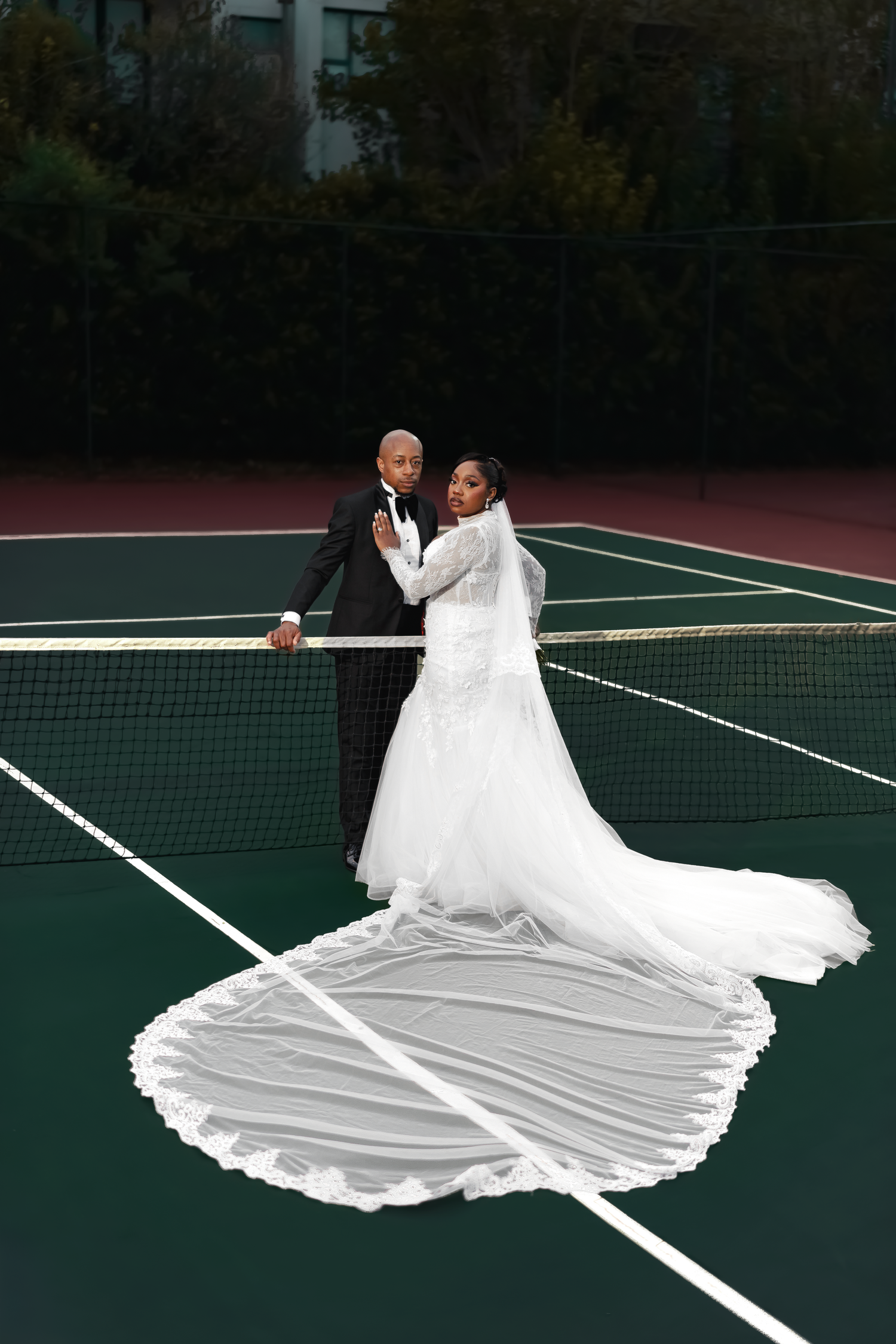 Bride and groom posing on a tennis court, bride wearing a stunning white gown with long flowing train at the net.