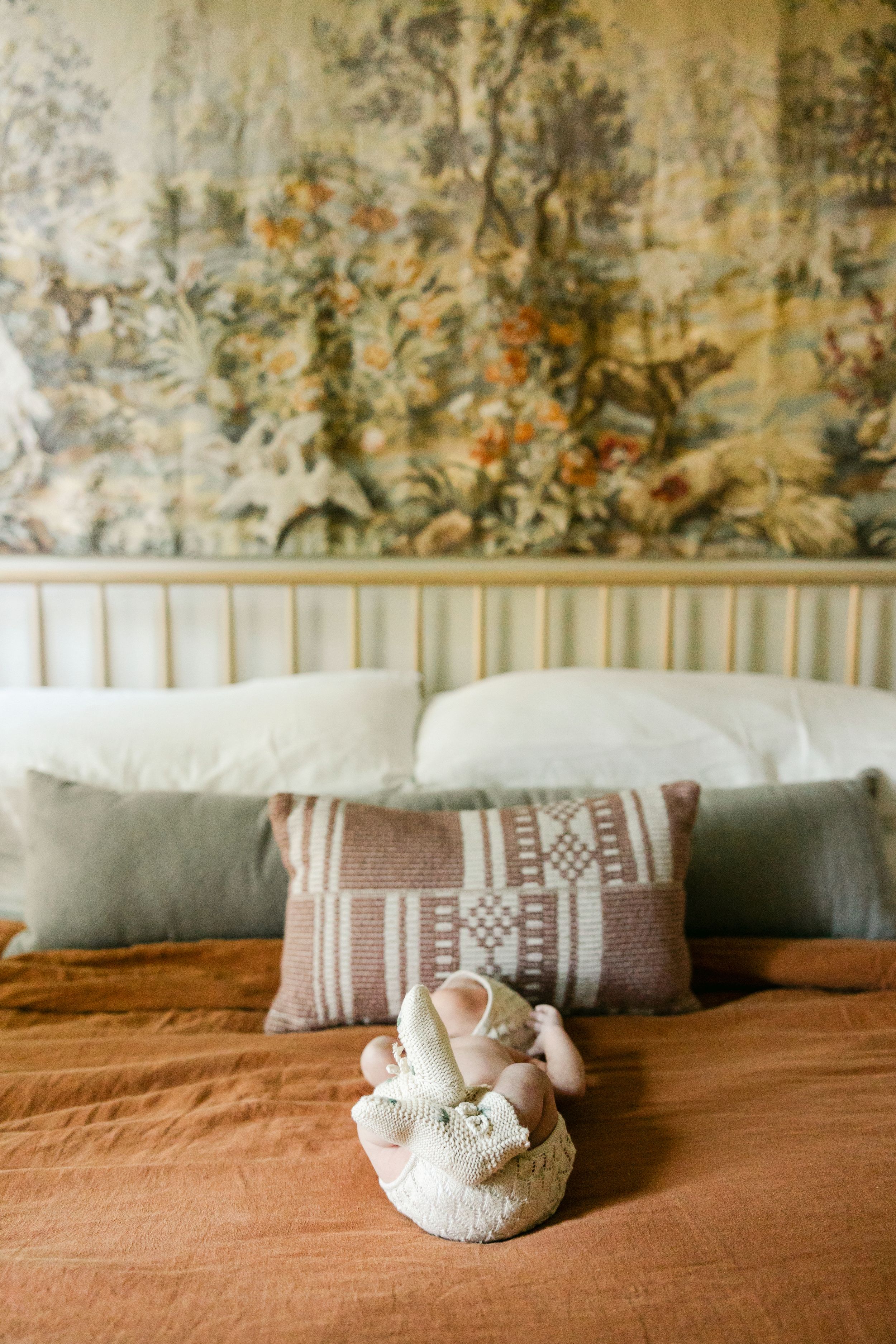 Elegant bed with rust linen, patterned pillows, white toy, and vintage floral tapestry wall hanging.