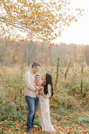A family of three poses outdoors in autumn, parents holding their baby girl among golden fall foliage.