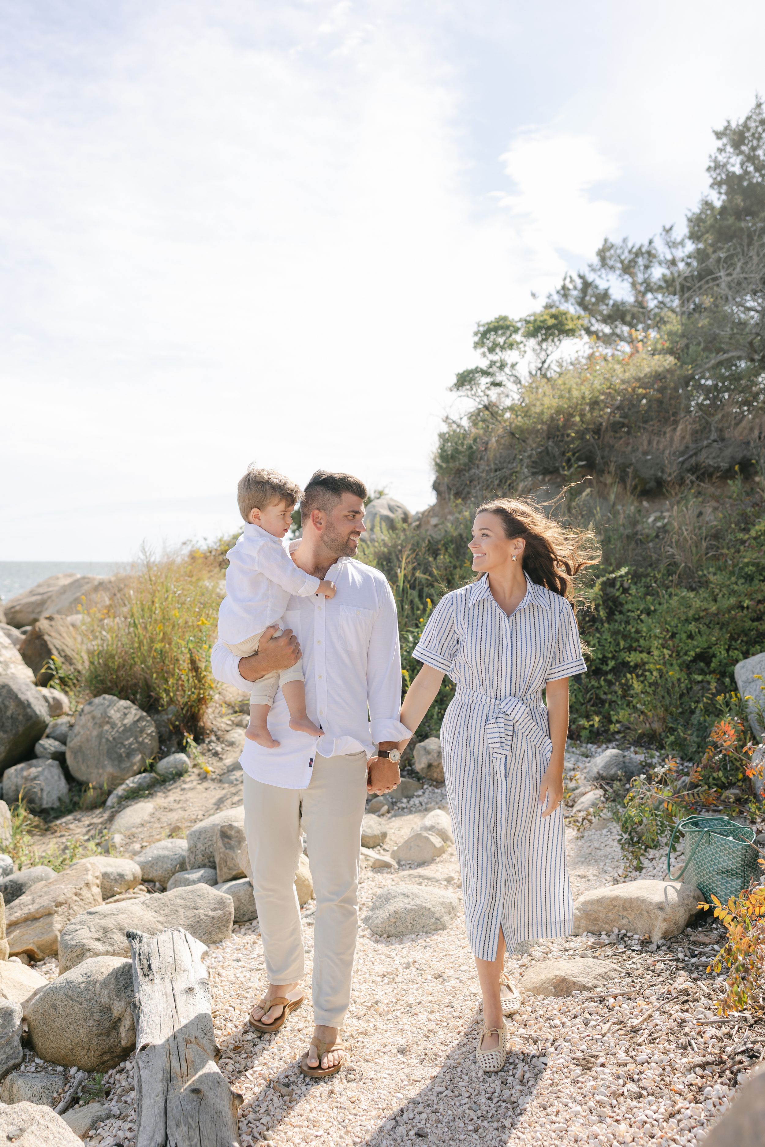 Happy family with toddler walking along a rocky coastal path on a sunny day near the ocean.