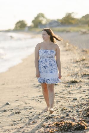 Young woman in floral strapless dress walking barefoot on sandy beach at golden hour.