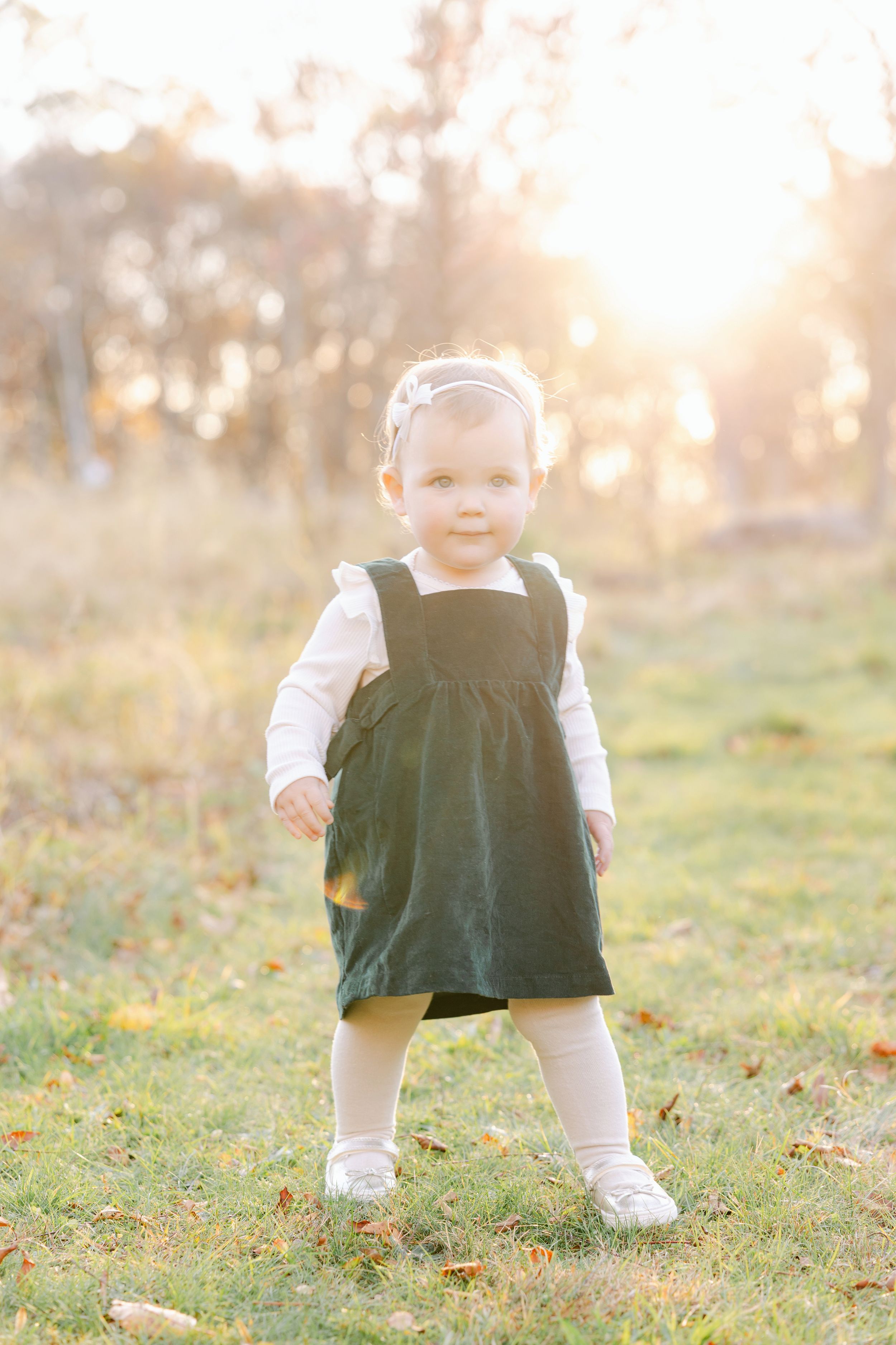 Toddler girl in dark green pinafore dress standing on grass in golden sunset light outdoors.