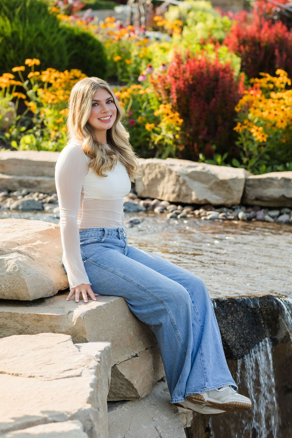Person in light sweater and flared jeans sits on stone wall surrounded by vibrant autumn flowers in garden setting.