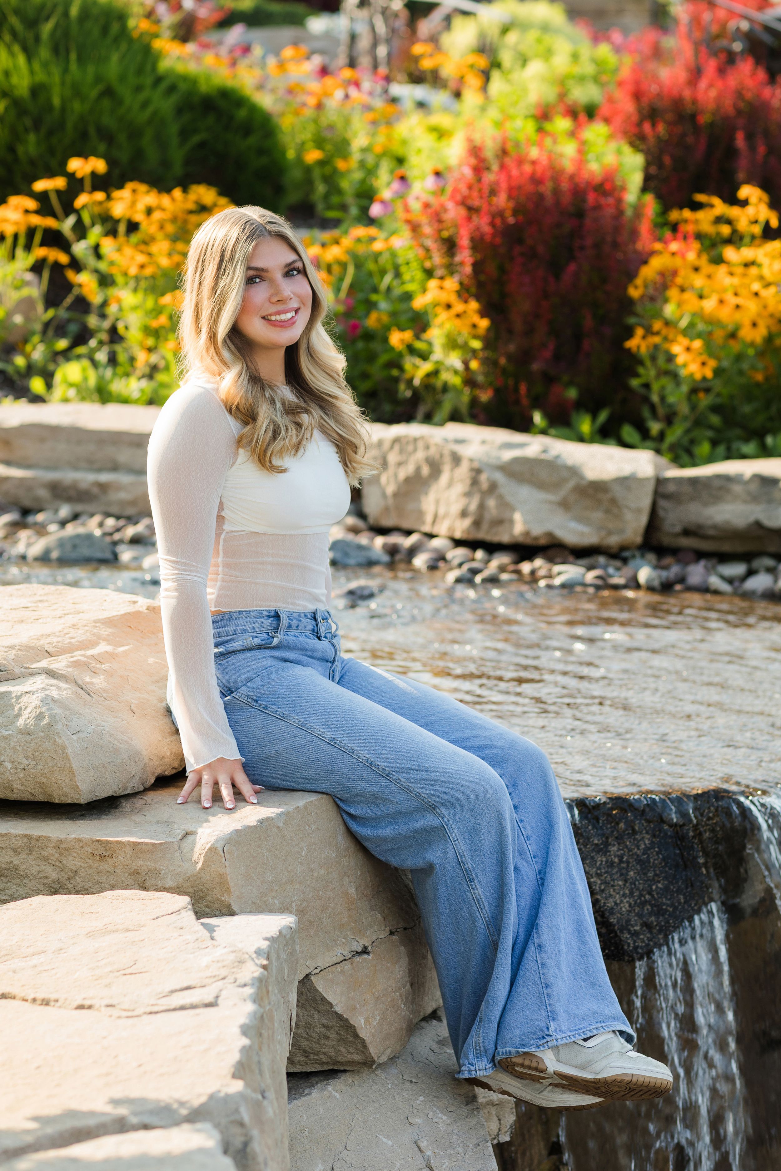 Person in light sweater and flared jeans sits on stone wall surrounded by vibrant autumn flowers in garden setting.