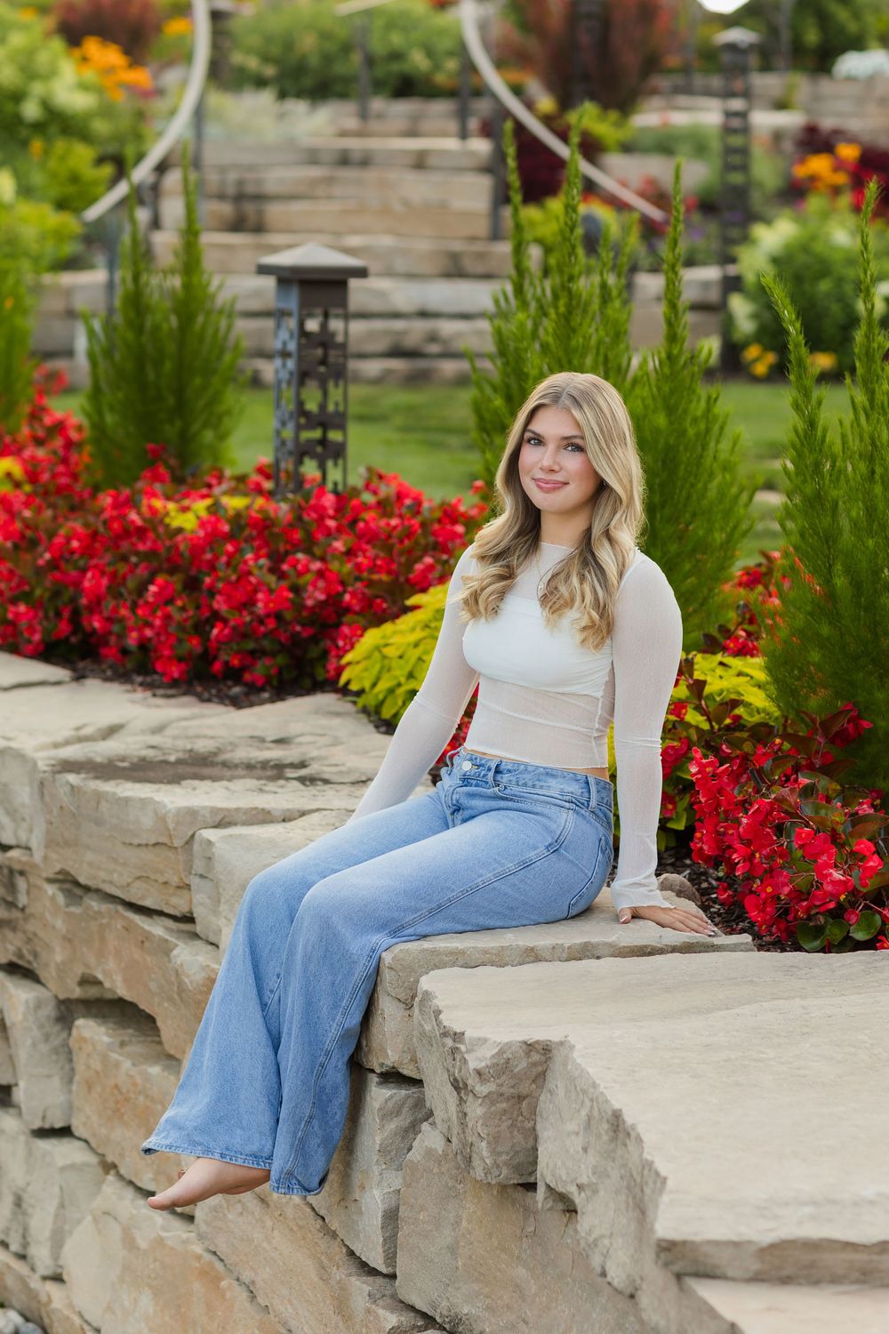 A person in a white top and light blue jeans sits casually on a stone wall surrounded by vibrant red flowers and greenery.