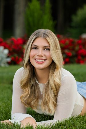 Portrait of someone with wavy blonde hair lying on grass with vibrant red flowers in the background.