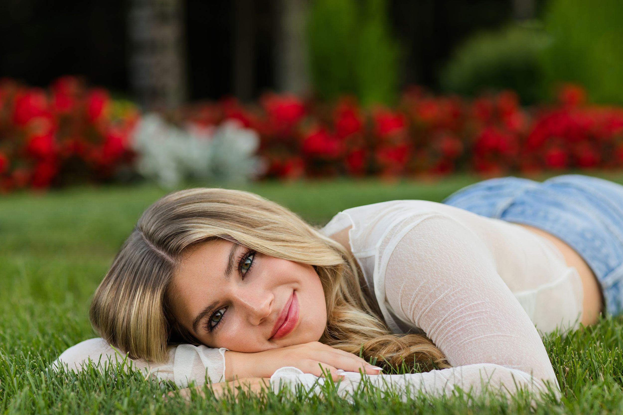 A person in a white dress lying on green grass with red flowers in the background.