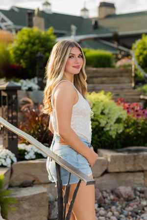 Woman in white top and denim shorts poses outdoors near garden steps and landscaping in summer setting.