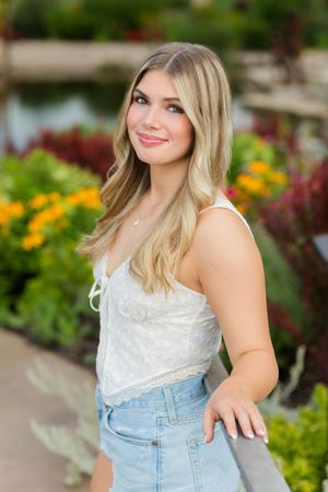 A person wearing a white lace top and denim overalls poses in a garden setting with colorful flowers in the background.