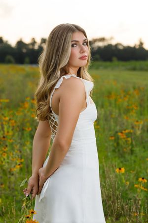 Woman in white summer dress stands gracefully among blooming wildflowers during sunset.