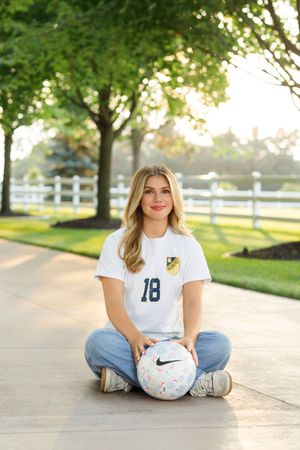 Casual outdoor senior photos with soccer ball on sidewalk near white fence.