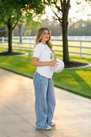 A person in jeans and white t-shirt poses with a volleyball in a sunny park setting with white fences.