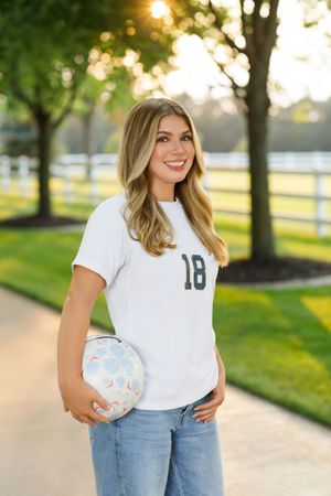 A person in a white t-shirt and jeans poses outdoors with a volleyball against a sunlit background with trees.