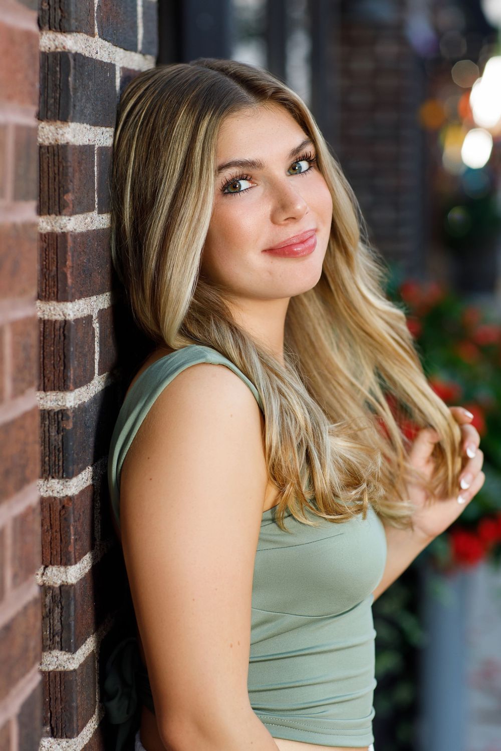 Portrait against brick wall wearing sage green tank top with styled blonde waves.