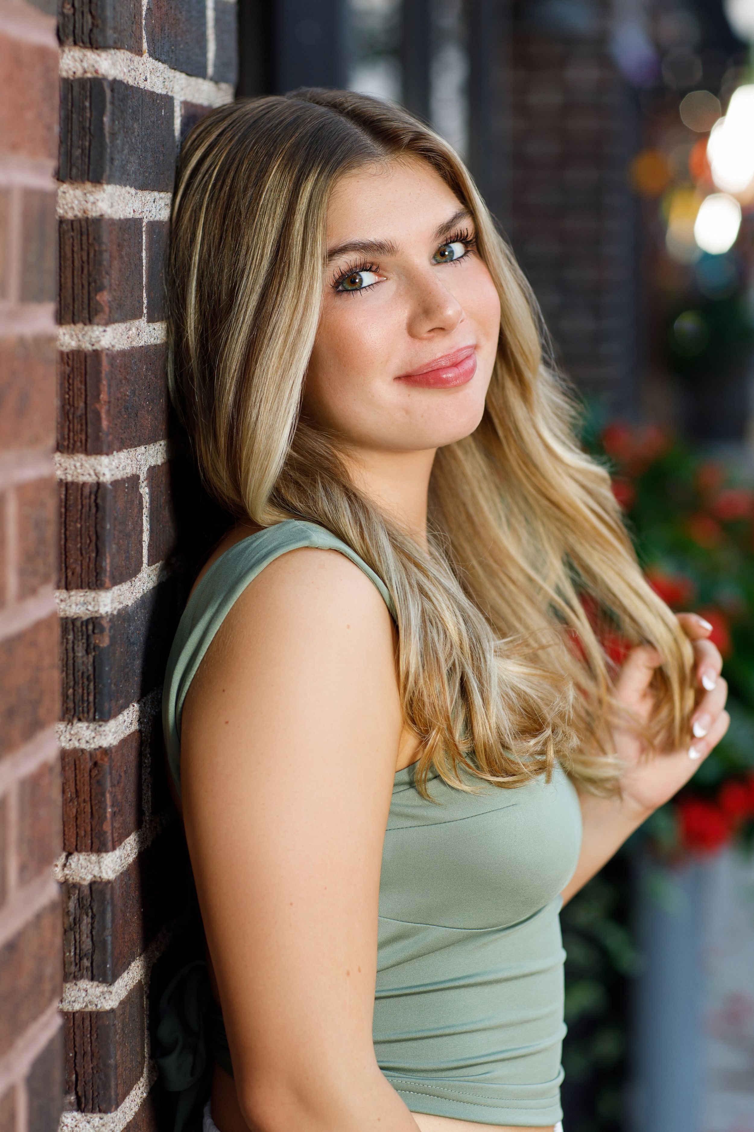 Portrait against brick wall wearing sage green tank top with styled blonde waves.