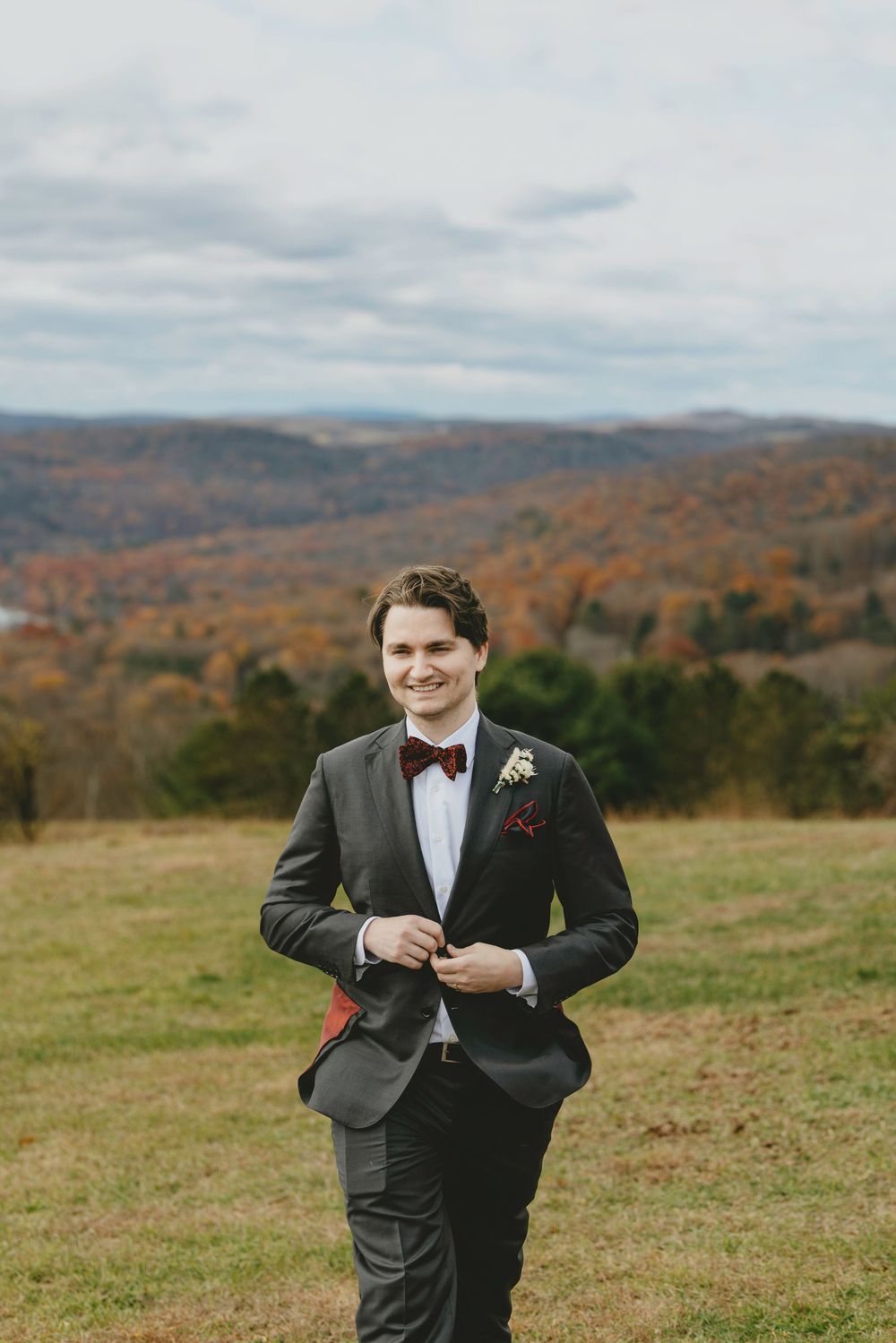 A groom in a black tuxedo poses in an open field with fall foliage and mountains in the background.
