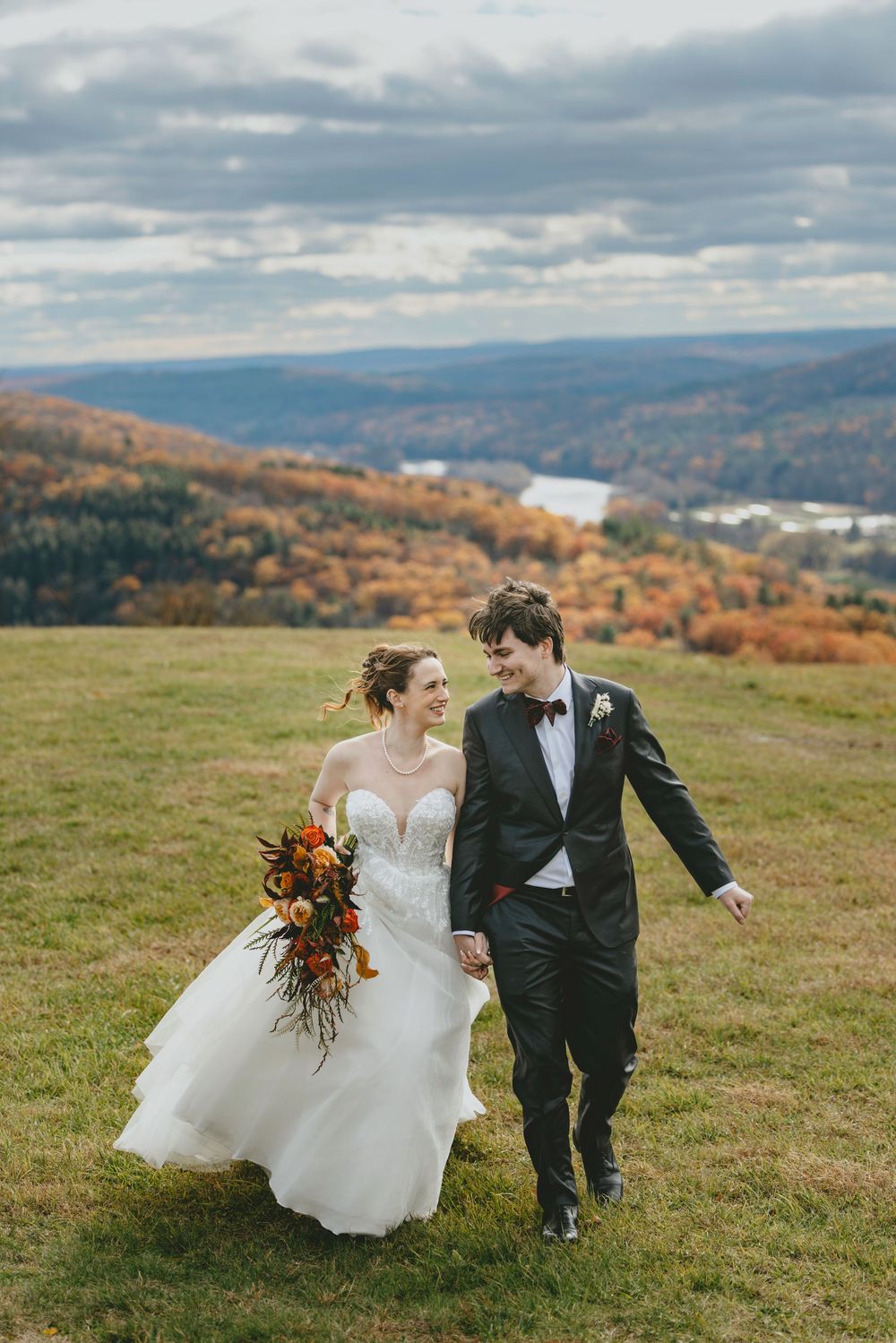 A couple in wedding attire walks hand in hand across a hilltop meadow with autumn mountains and a lake in the background.