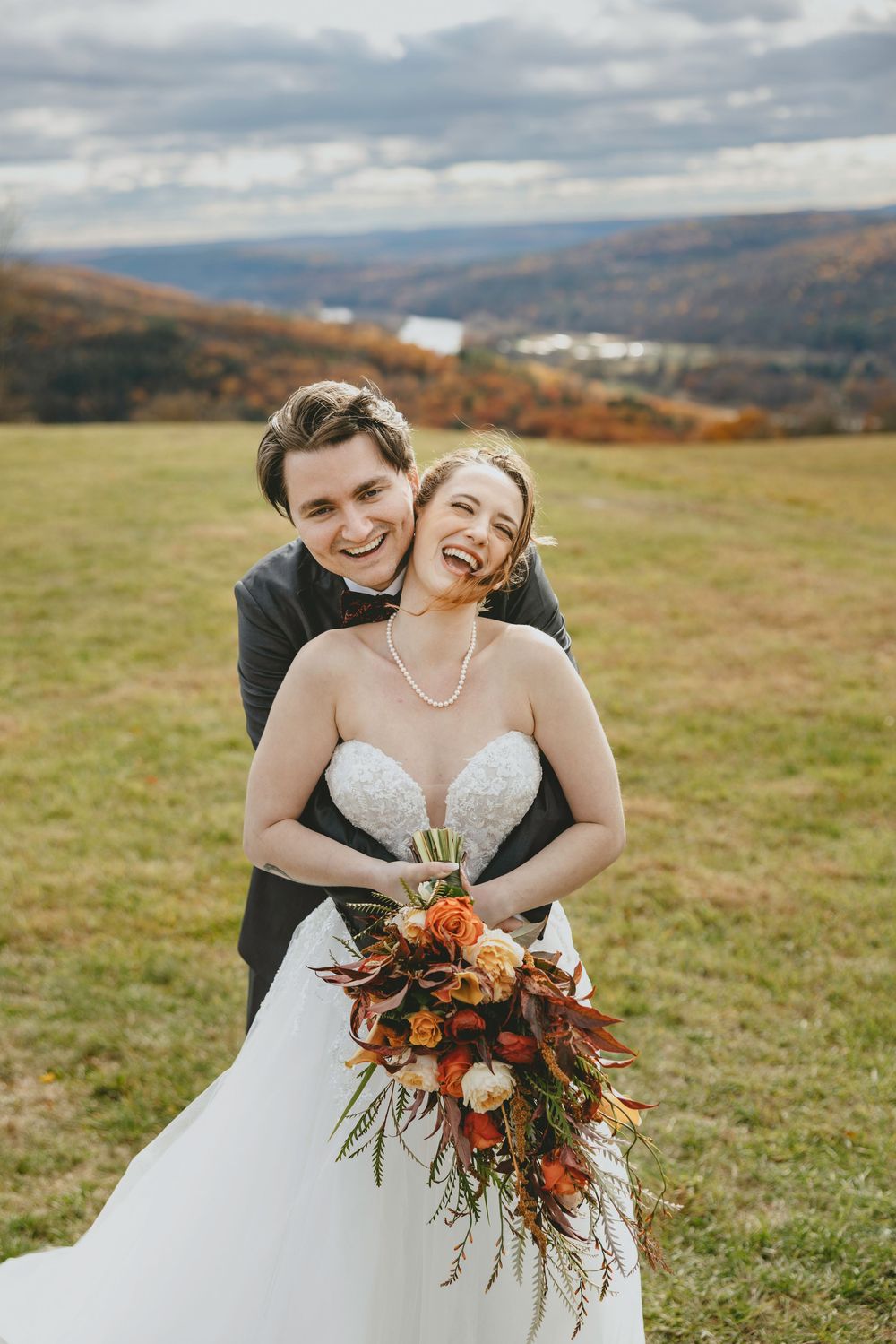 Two brides share a joyful moment together on a hilltop with autumn mountains in the background.