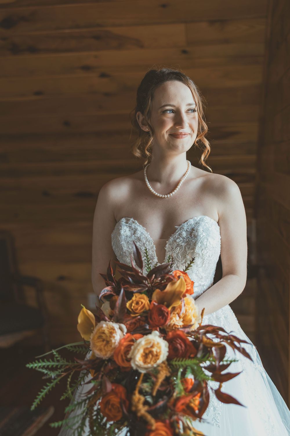 Bride in white strapless gown holds vibrant orange and peach floral bouquet against rustic wooden backdrop.