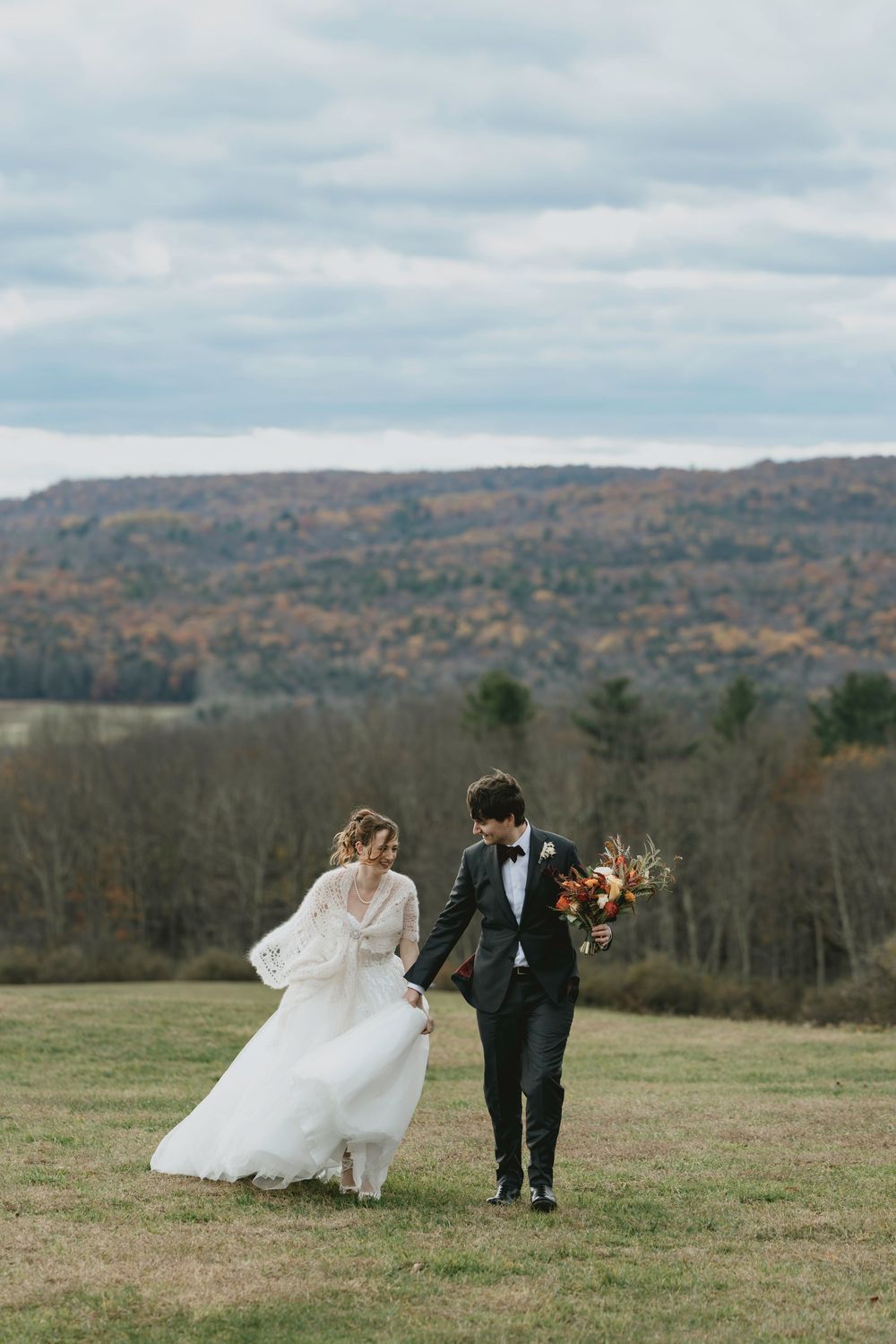 A couple in wedding attire walks across a hilltop meadow with autumn foliage in the background on a cloudy day.